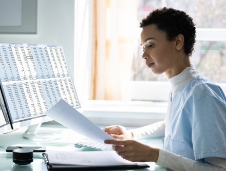 Woman in scrubs working on paperwork at a desk with a computer and spreadsheet